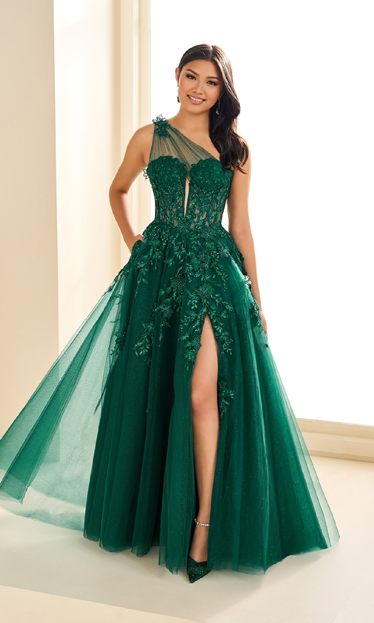 Woman wearing the Ellie Wilde EW36207 long prom dress featuring a sleeveless, dark green design with floral appliques, a sheer corset bodice, and a high slit, standing indoors and smiling at the camera.